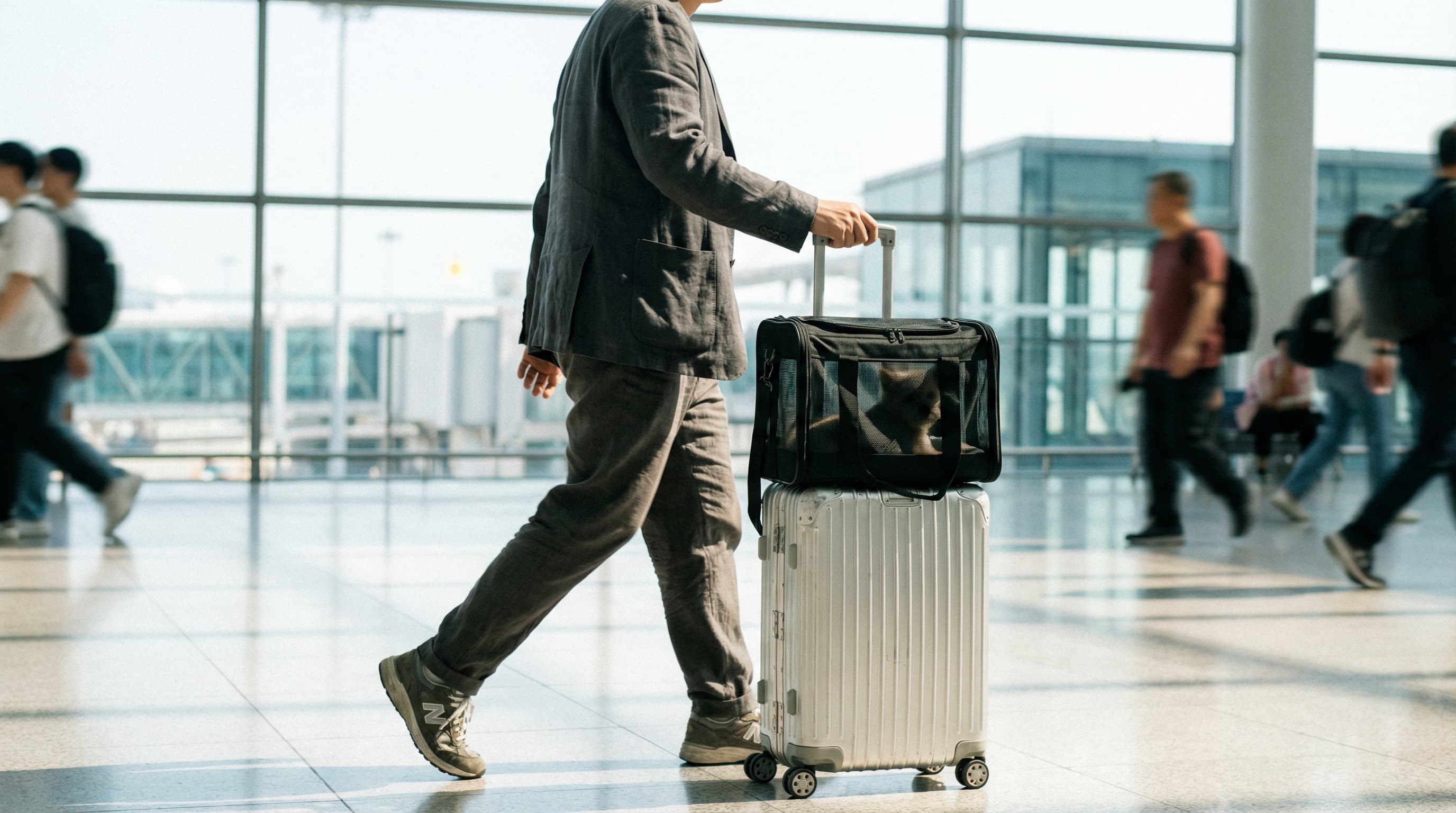A traveler pulling a silver rolling suitcase with a sleek black pet carrier secured to the handle via a trolley sleeve.